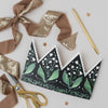 Decorative paper crown with green leaf design, gold scissors, and ribbons on a light gray background