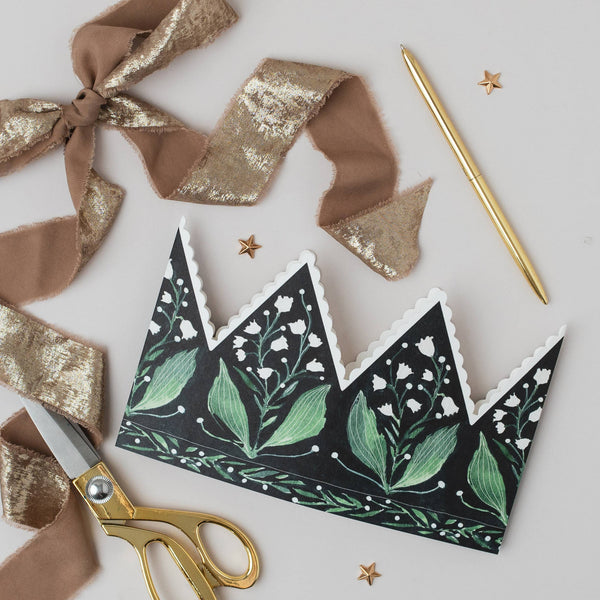 Decorative paper crown with green leaf design, gold scissors, and ribbons on a light gray background