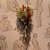 Dried flowers and herbs on a beige background with black line drawings of nude figures.